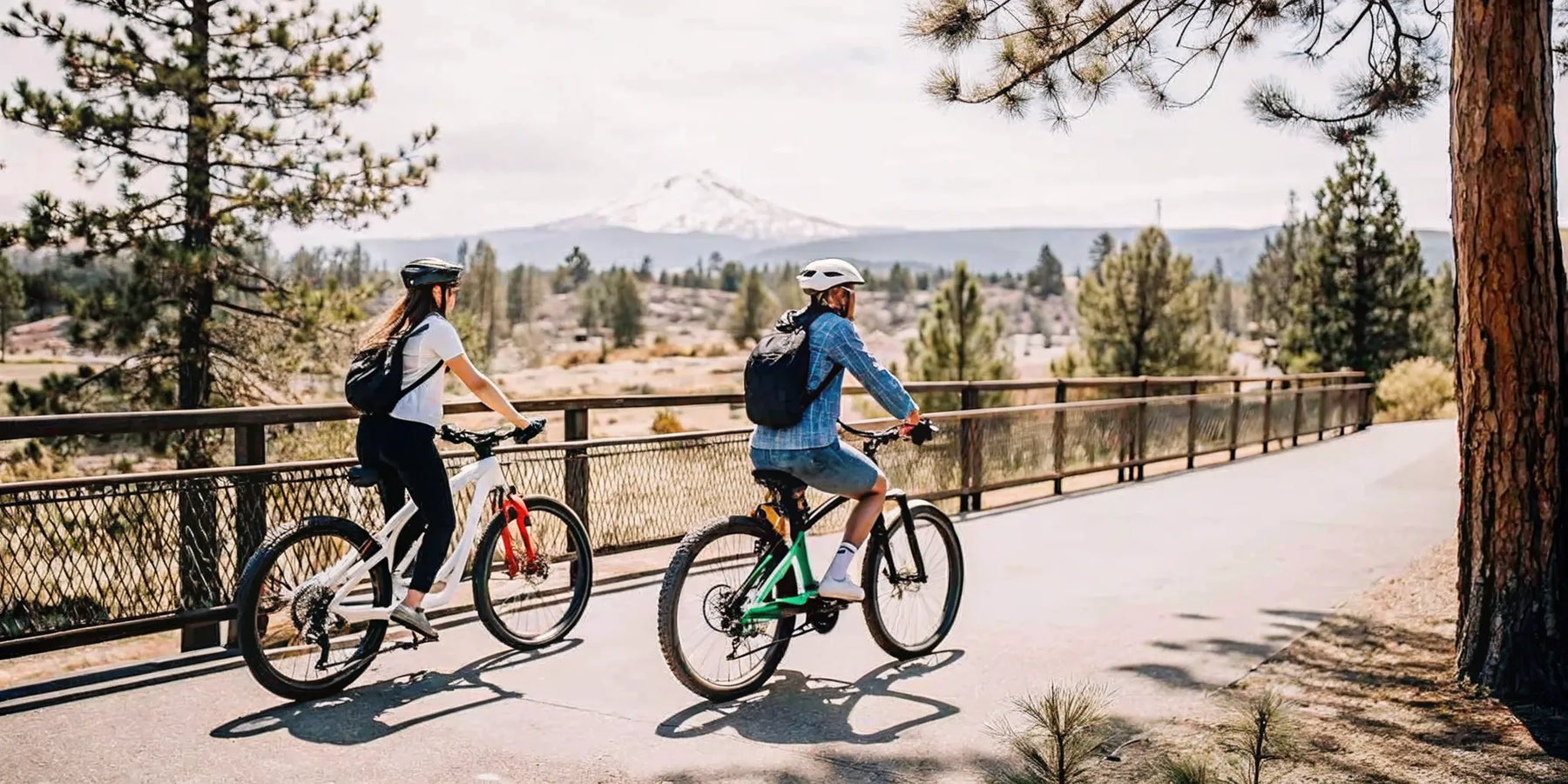 Two cyclists riding electric bikes along the Deschutes River Trail in Bend, Oregon on a sunny day with Cascade Mountains in the background